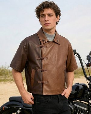 Front lifestyle model view of a sample custom men's two-tone short-sleeve leather shirt jacket displaying toggle loop closures, asymmetric brown panels, and a motorcycle backdrop.