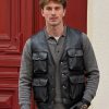 Front lifestyle model view of a sample custom men's black leather multi-pocket utility vest featuring four front cargo pockets and brass snap buttons over a grey polo sweater.